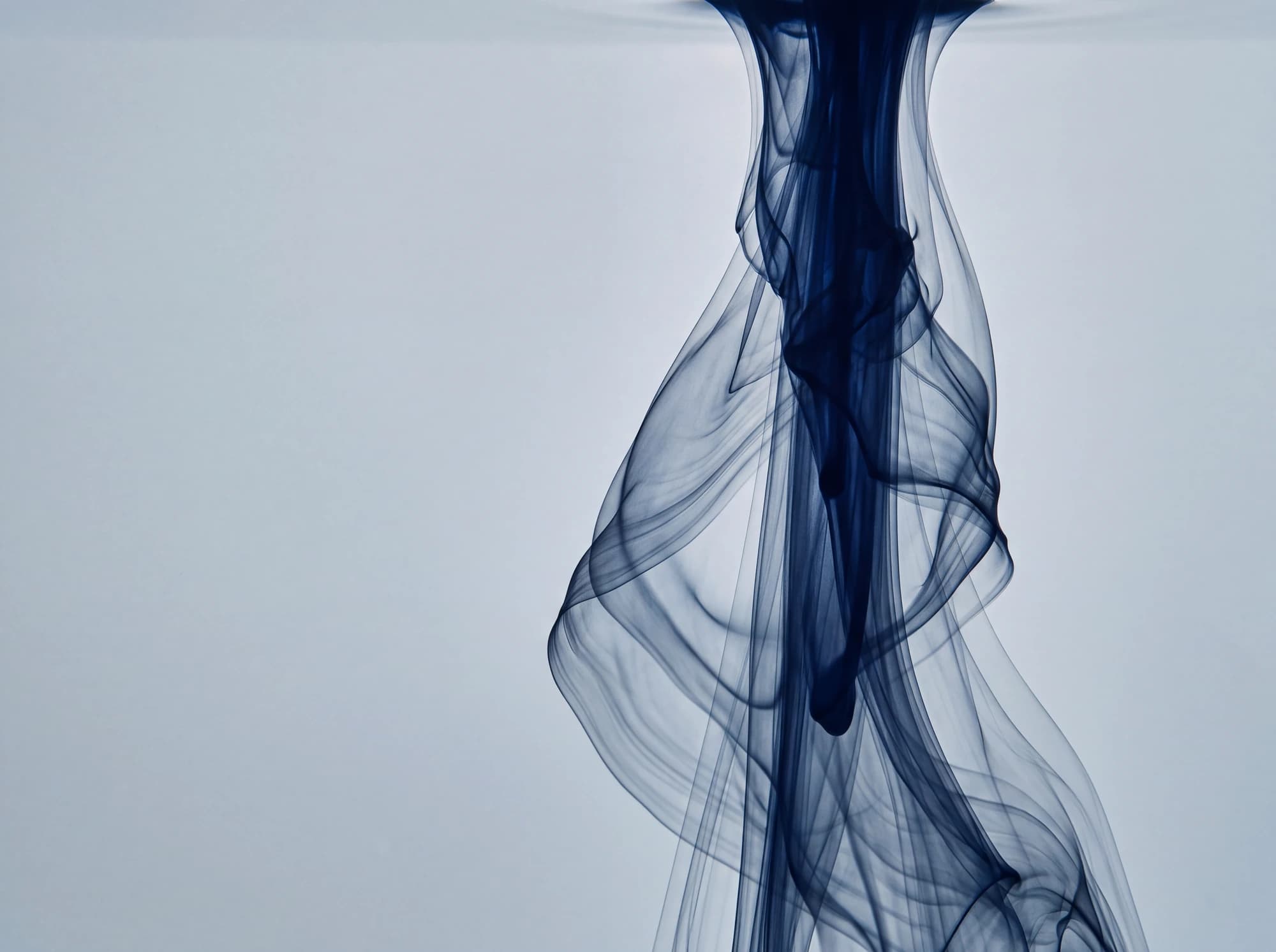 A single drop of navy ink unfurling into overlapping veils in still pale water
